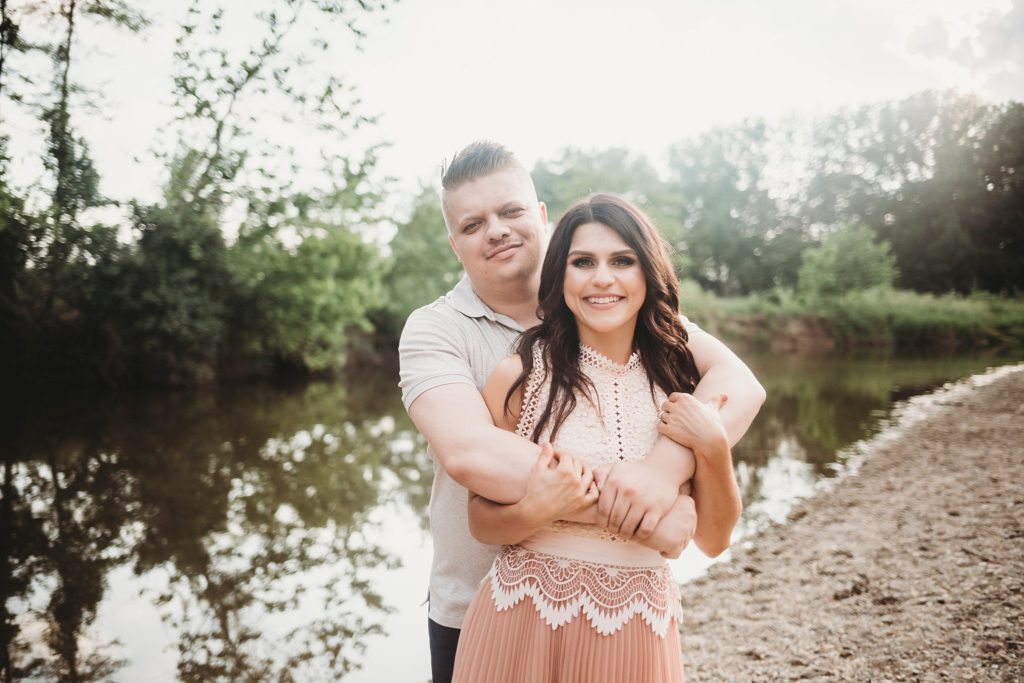 Fairfield Lakes Park Engagement Session- Man hugging woman from behind while smiling near water in Lafayette Indiana at Fairfield lakes park.