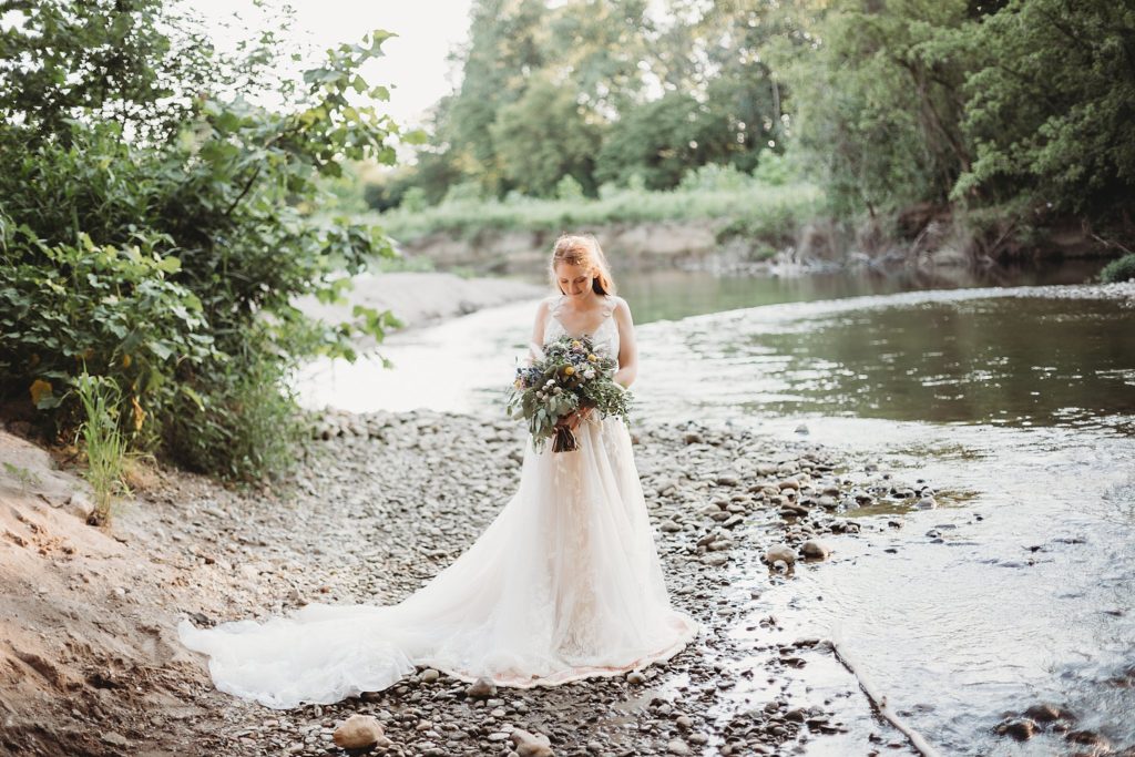 Indiana Elopement// Morgan + Kyle - Bride stands alone near the water looking at her bouquet peacefully.