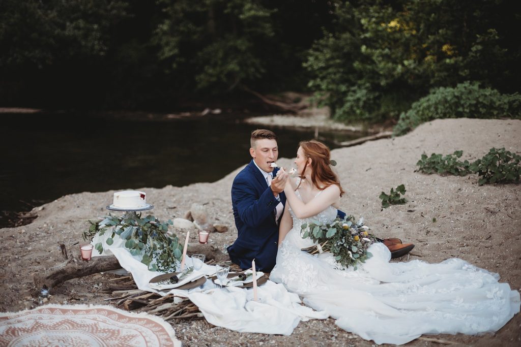 Indiana Elopement// Morgan + Kyle -  Couple sits in the sand together and feed each other bites of wedding cake.