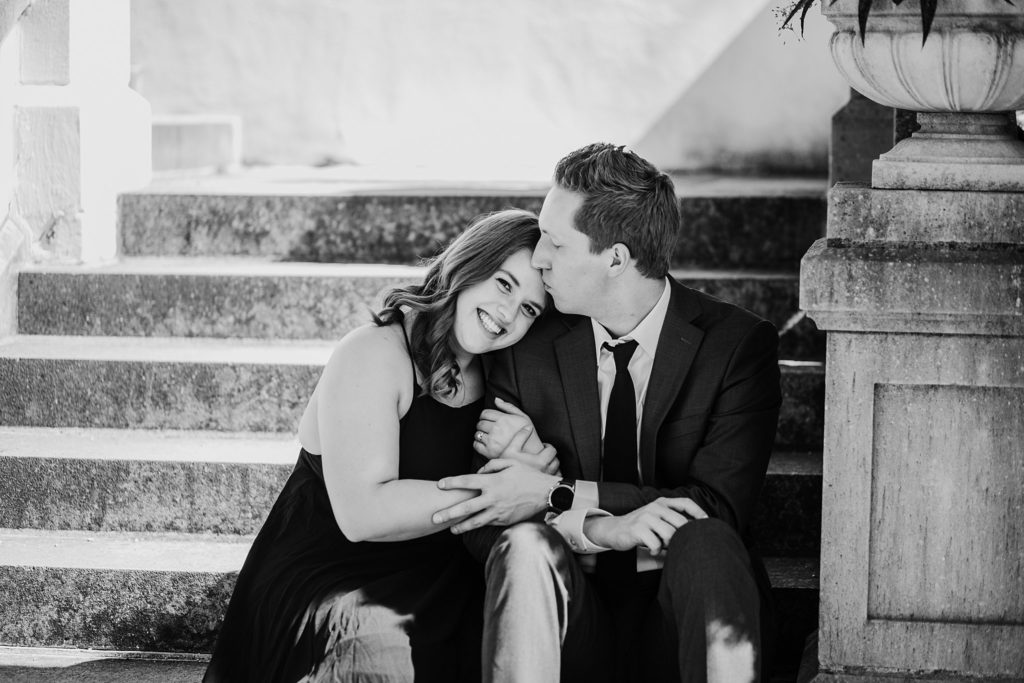 Newfields Engagement Session// Michelle + Adam - Couple sits together on the stairs. Man kisses woman's forehead as woman smiles at the camera.