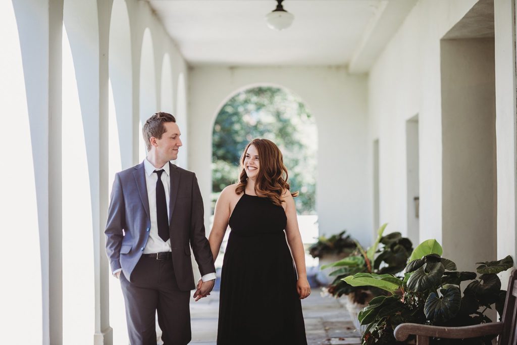 Newfields Engagement Session// Michelle + Adam - Man and woman walk together and smile at each other under a covered patio.