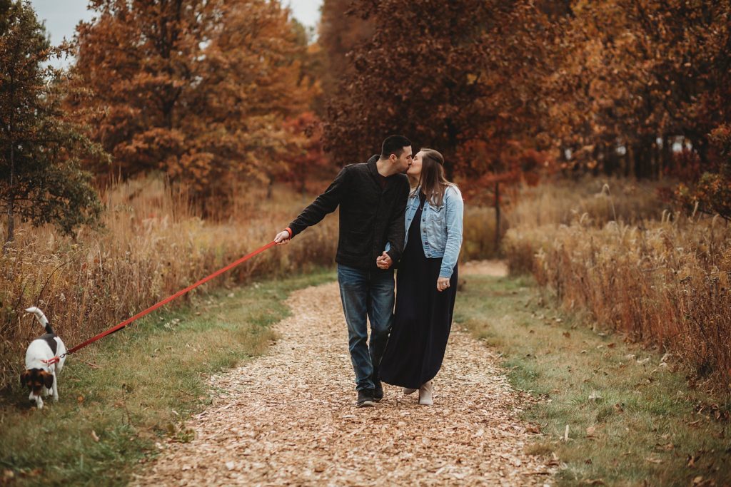 man and woman walk their dog in a park for their engagement pictures 