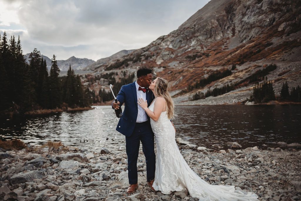 groom shaking a champagne bottle while kissing his bride at Blue Lakes in Breckenridge Colorado. 