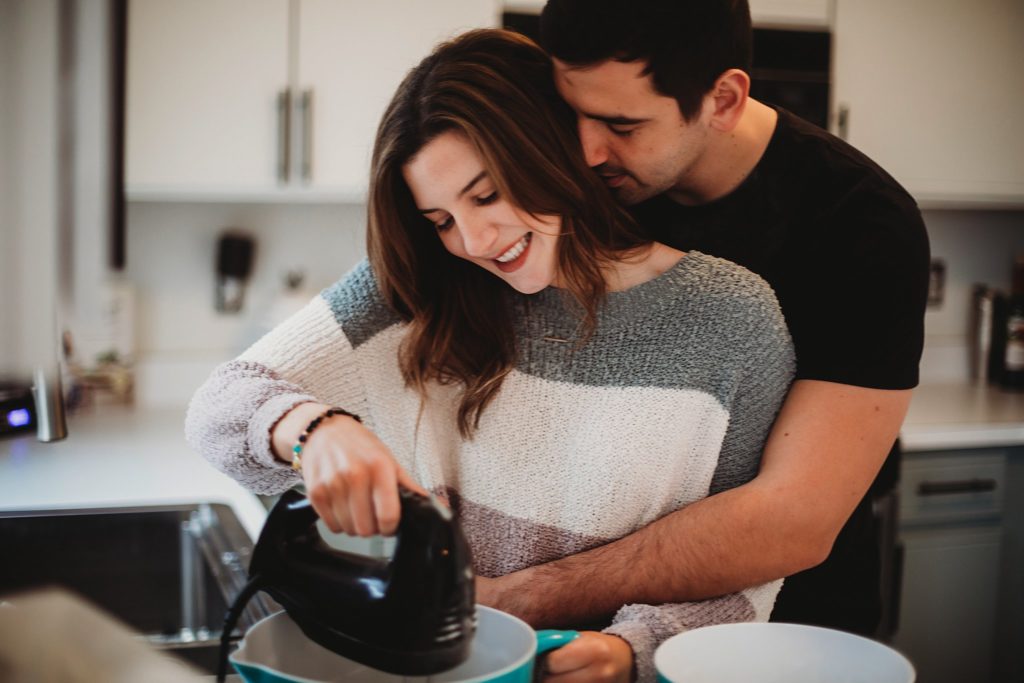 Man wrapping his hands around a woman and nuzzling in her neck while she mixes batter during their crown point indiana engagement session