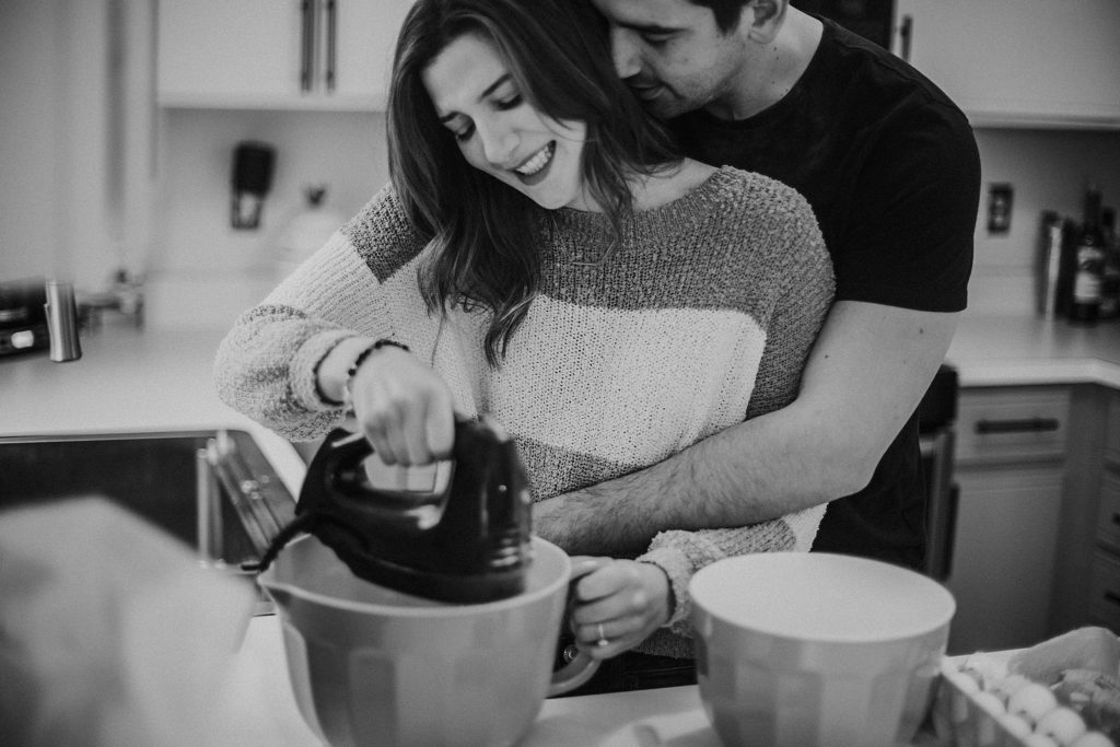 Man holding around woman's waist while she is mixing flour in a bowl at their Crown Point Indiana Engagement Session