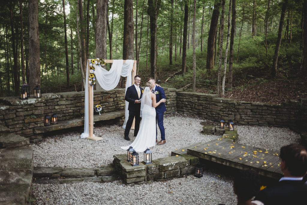 officiant standing in front of backdrop watching bride and groom kiss in front of guests