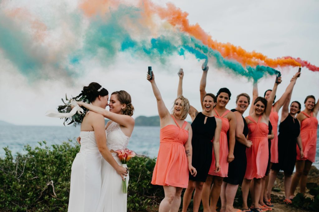 LGBTQ+ Friendly Honeymoon Destinations- two wives looking at each other while rainbow smoke bombs are in the background with wedding party at St. Thomas, Virgin Islands Wedding.