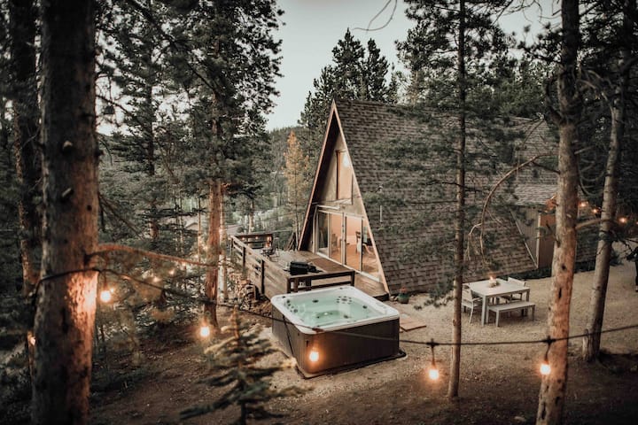 Best Colorado A-Frames- An A-frame cabin in Breckenrdige, Colorado is surrounded by trees. Also pictured is a hot tub, picnic table, and lights. 