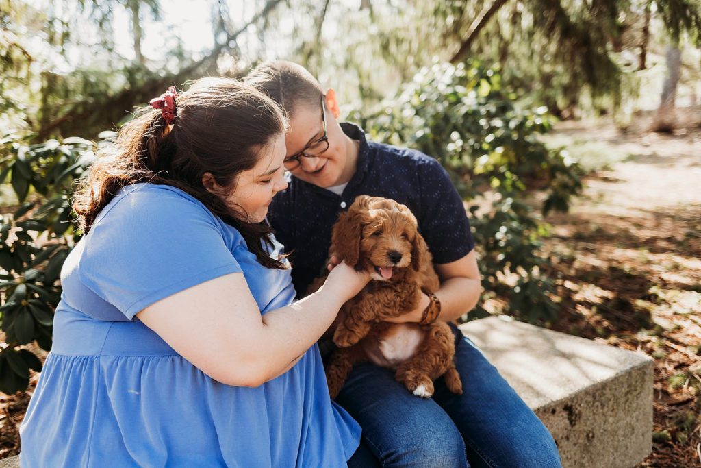 couple sitting on a bench in the woods while holding golden puppy and smiling