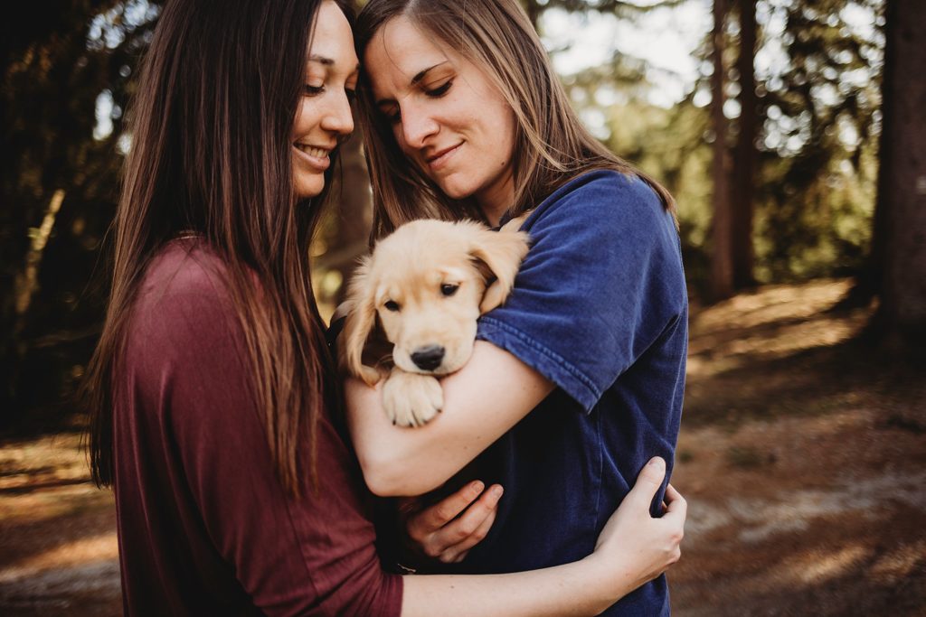 girl in red shirt holding girl in blue while holding puppy in arms gazing down at it.