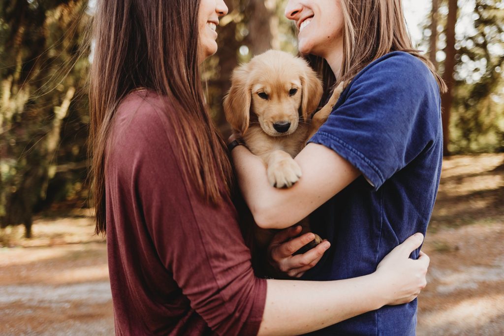 girl in red shirt holding girl in blue while holding puppy in arms smiling at eachother