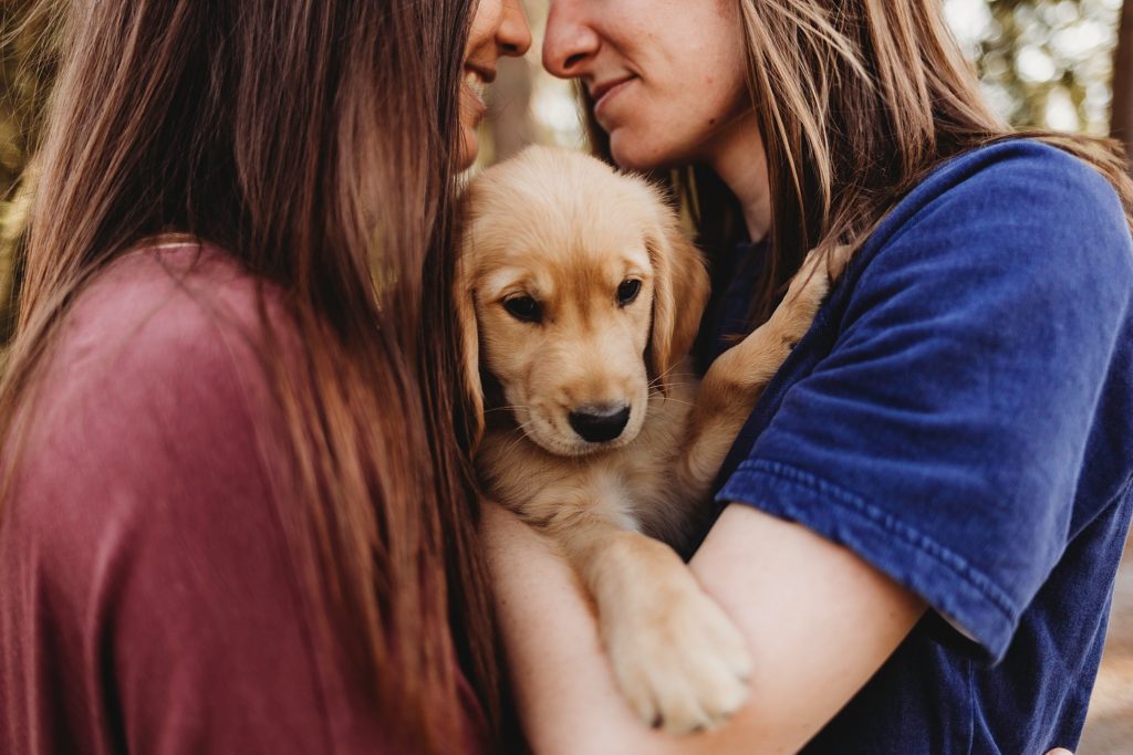 girl in red shirt holding girl in blue while holding puppy in arms gazing down at each other.