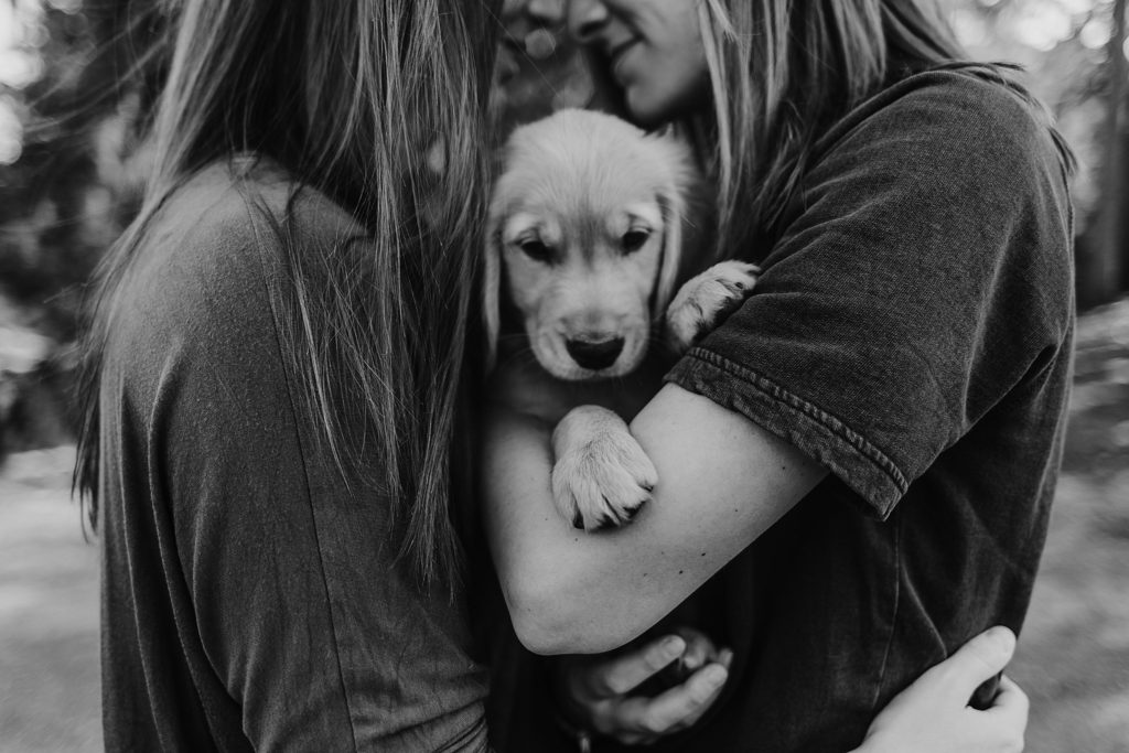black and white picture of girl in red shirt holding girl in blue while holding puppy in arms gazing down at each other.