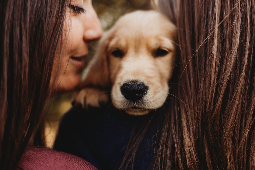 Woman in blue shirt holding puppy over shoulder with back to the camera while girl in red shirt prepares to kiss the puppy's head at Purdue horticulture park.