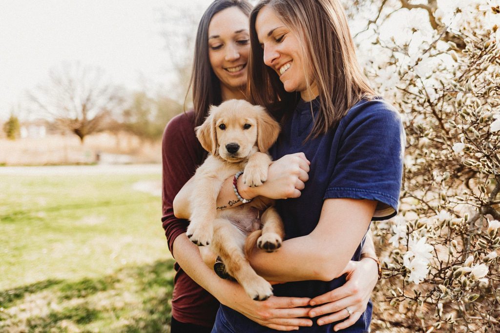 girl in red shirt holding girl in blue while holding puppy in arms gazing down at it and white and brown bush in background