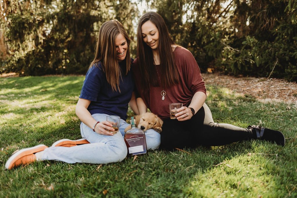 Two women sitting in the grass at Purdue horticulture park while touching glasses and a puppy laying between them and gazing down at it.