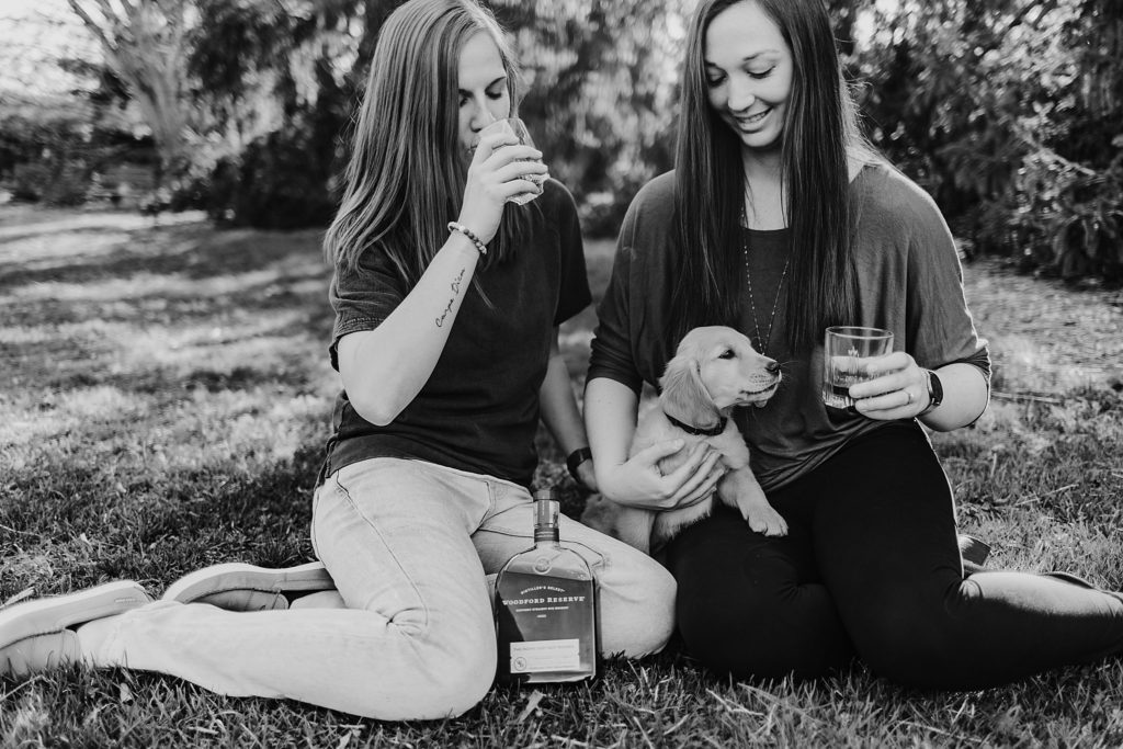 black and white picture of couple sitting in the grass drinking out of glasses and puppy showing interest in the glasses.