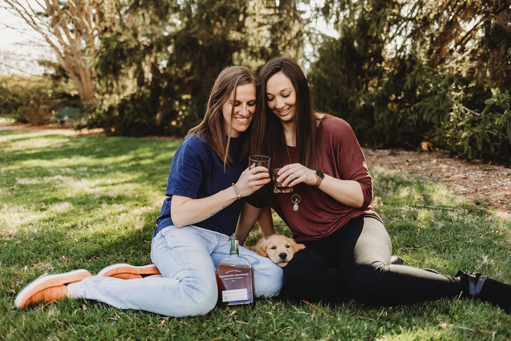 Two women sitting in the grass at Purdue horticulture park while touching glasses and a puppy laying between them.