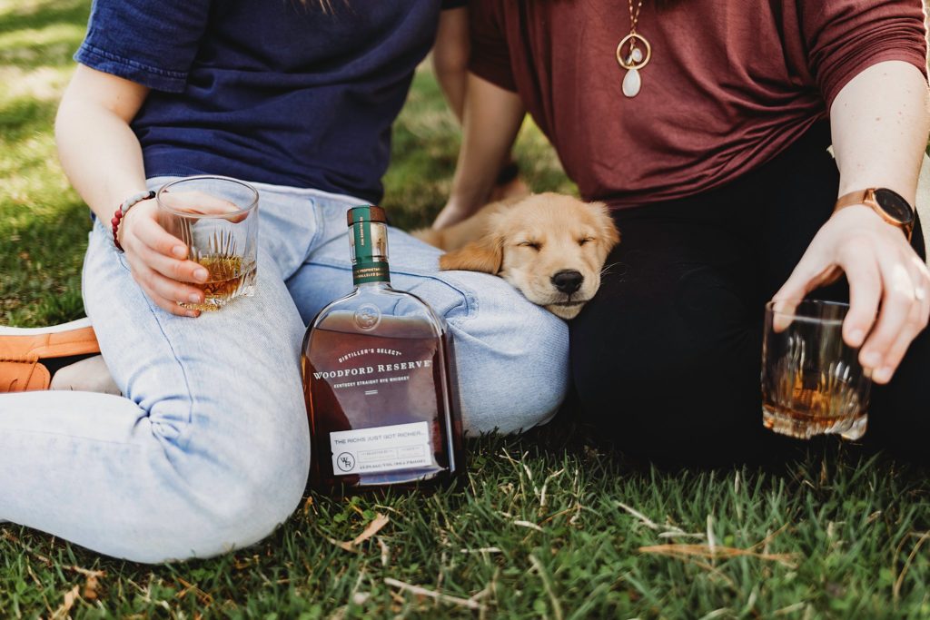 Two women sitting in the grass at Purdue horticulture park while showing whiskey glasses and a puppy laying between them.