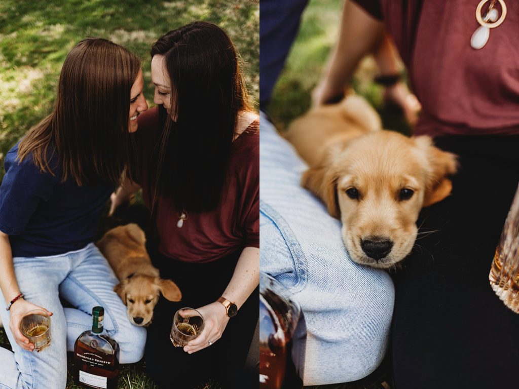 Two women sitting in the grass at Purdue horticulture park while showing whiskey glasses and a puppy laying between them with the camera angle pointing down