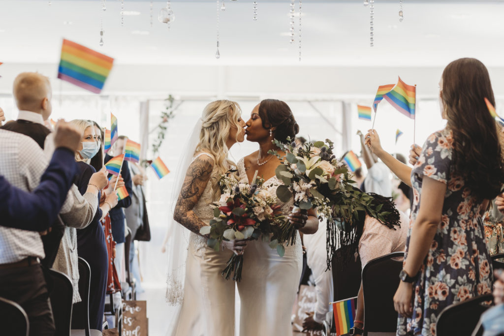 Two women kissing in the middle of isle while guests hold pride flags