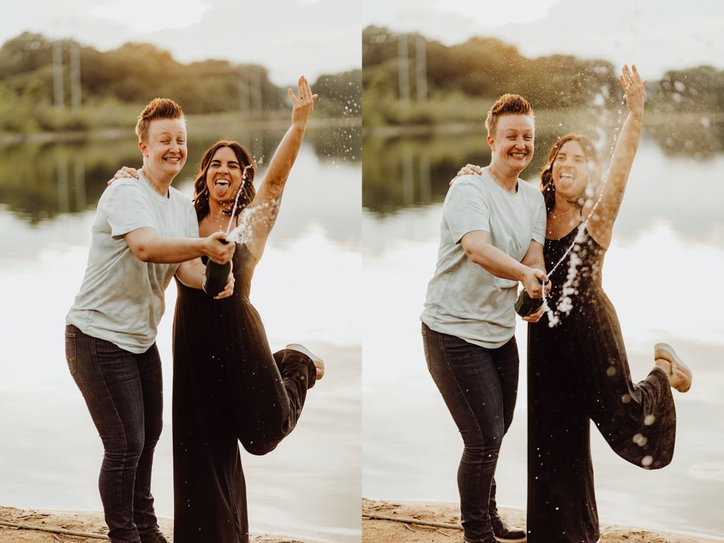 LGBTQ engagement session-two women holding hands in front of a lake while shaking a bottle of champagne in west lafayette, indiana
