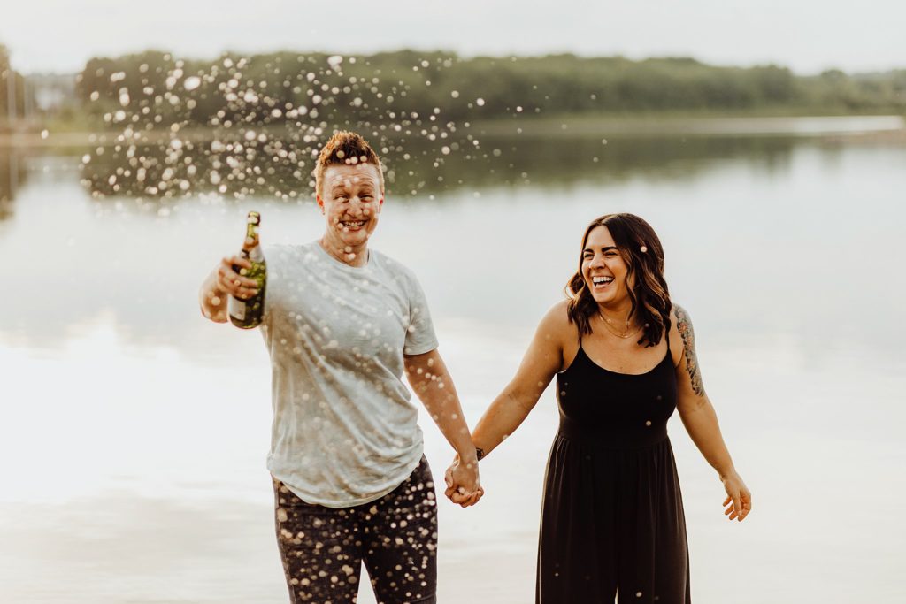 LGBTQ engagement session-two women holding hands in front of a lake while shaking a bottle of champagne in west lafayette, indiana