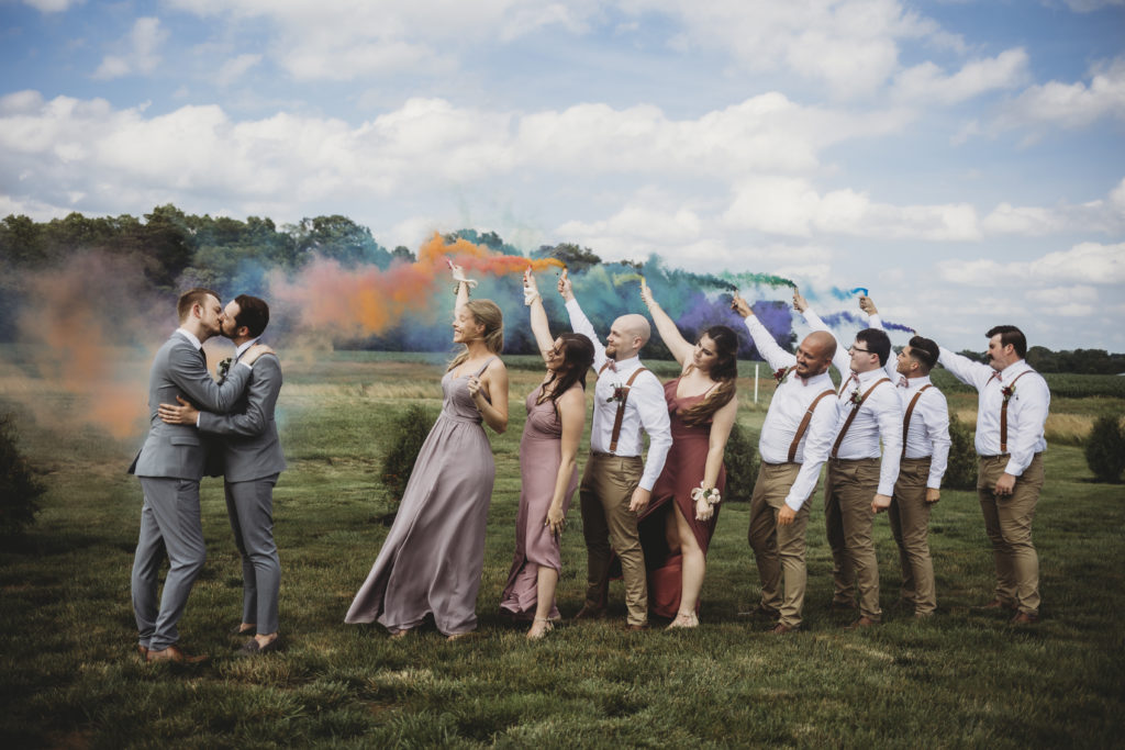 man and man kissing while wedding party holds up smoke bombs at their New Journey Farms Wedding in Lafayette, Indiana
