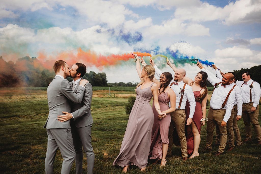 husband and husband kissing outside while their wedding party holds up rainbow colored smoke bombs at their LGBTQ friendly wedding venue 