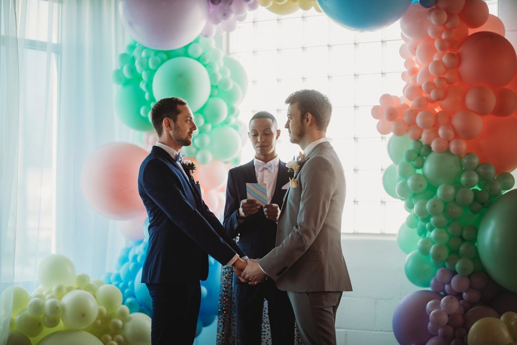 two men holding hands in front of a window that has a rainbow colored balloon arch with some of the best Indiana LGBTQ Wedding Vendors