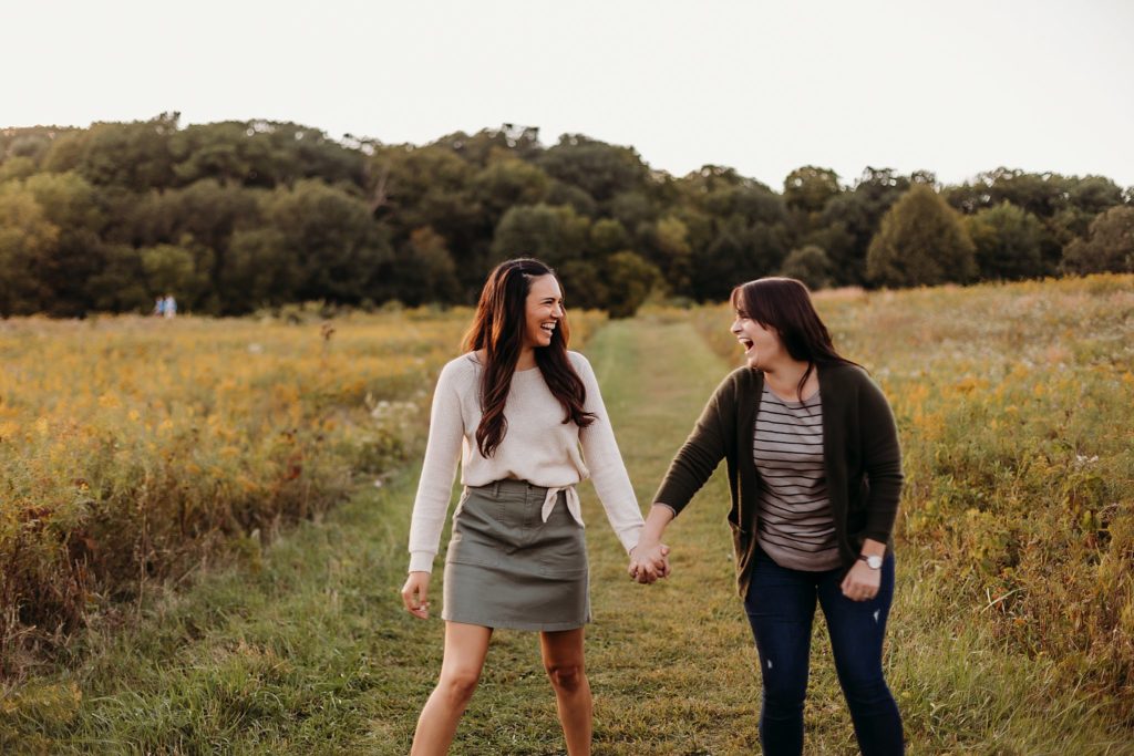 two women walk holding hands through a field of yellow flowers during their Chicago LGBTQ Couples Session