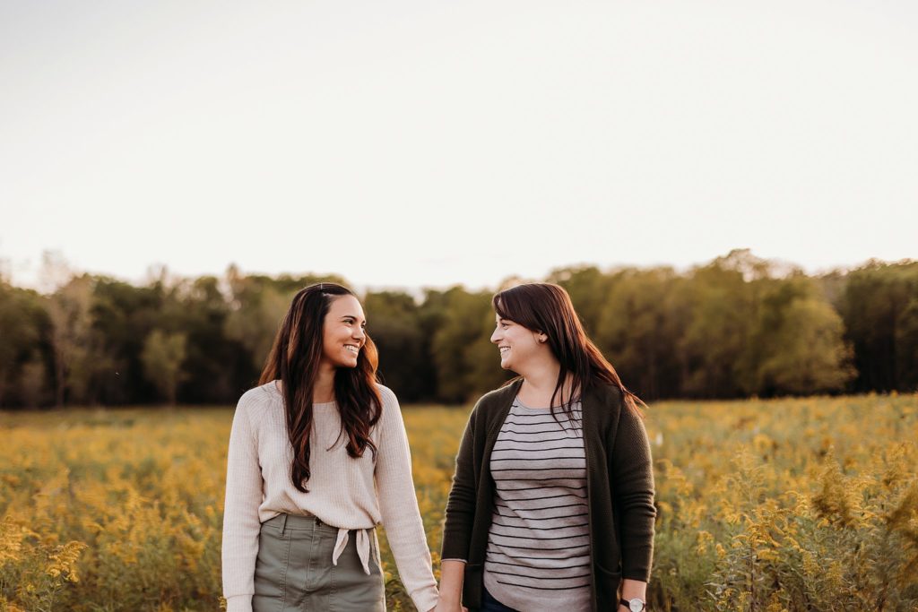 two women walk holding hands through a field of yellow flowers during their Chicago LGBTQ Couples Session