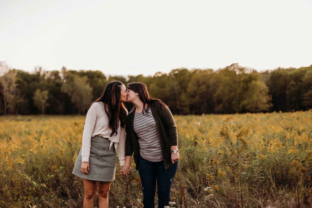 two women walk holding hands and kissing through a field of yellow flowers during their Chicago LGBTQ Couples Session