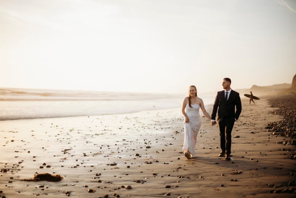 couple walking hand in hand along San Onofre State Beach while a surfer walks with a surfing board behind them at their California Elopement