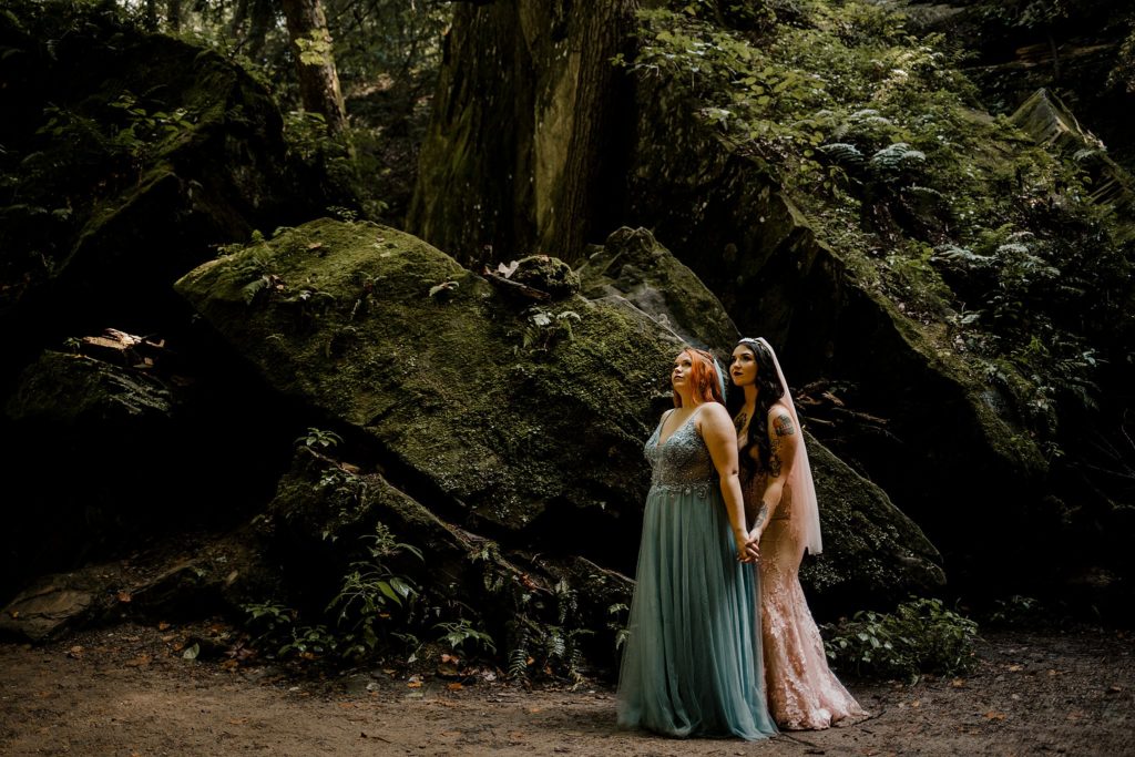 Two women hold onto one another while looking above them at the forest and trees while eloping in indiana