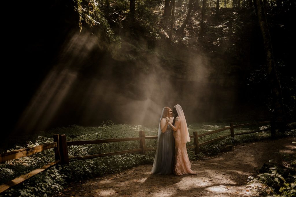 two women eloping in indiana, light shining in from the lush trees as they hold hands and smile at one another in Turkey Run State Park in Indiana