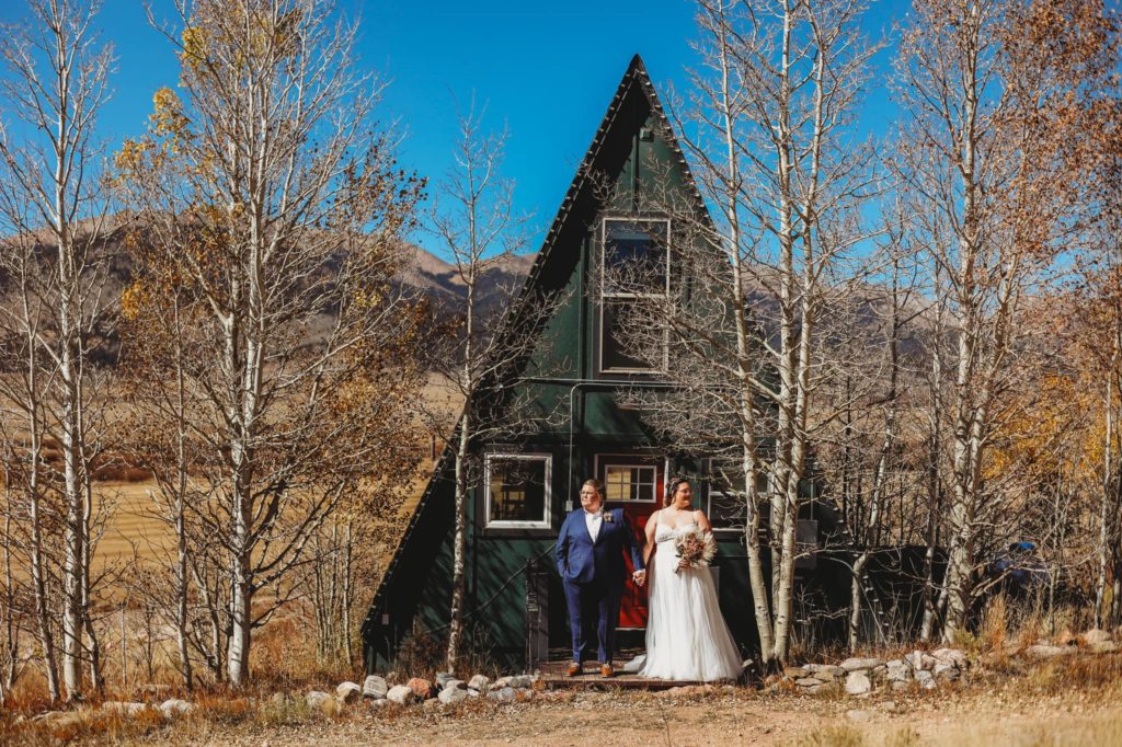 Samantha Mitchell photography shares one of the best lgbtq wedding photos- An lgbtq couple stand holding hands facing out towards the mountains in front of an A Frame Cabin in Jefferson, Colorado.